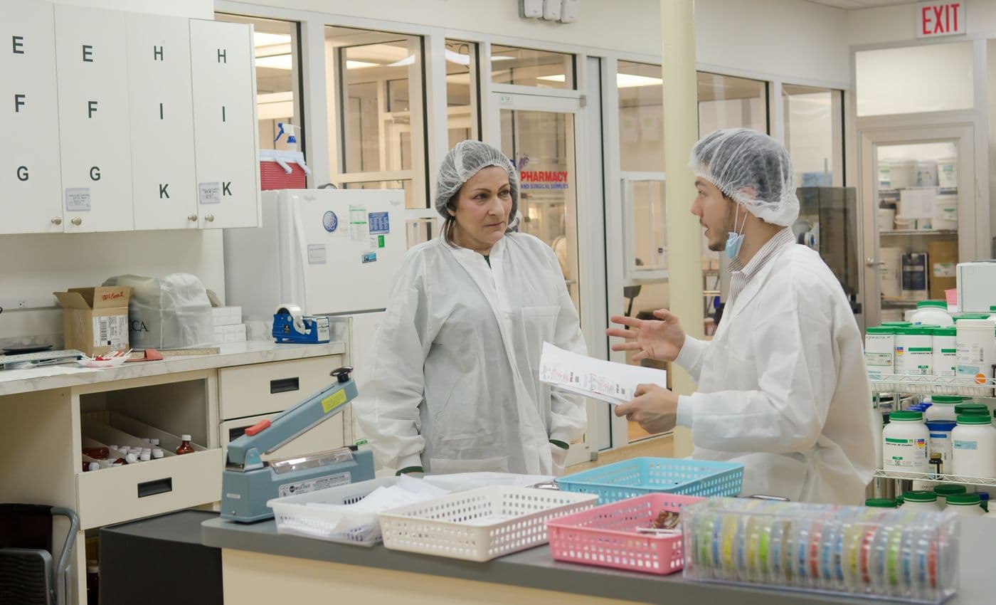 Two technicians in lab coats and caps review paperwork in a preparation lab.