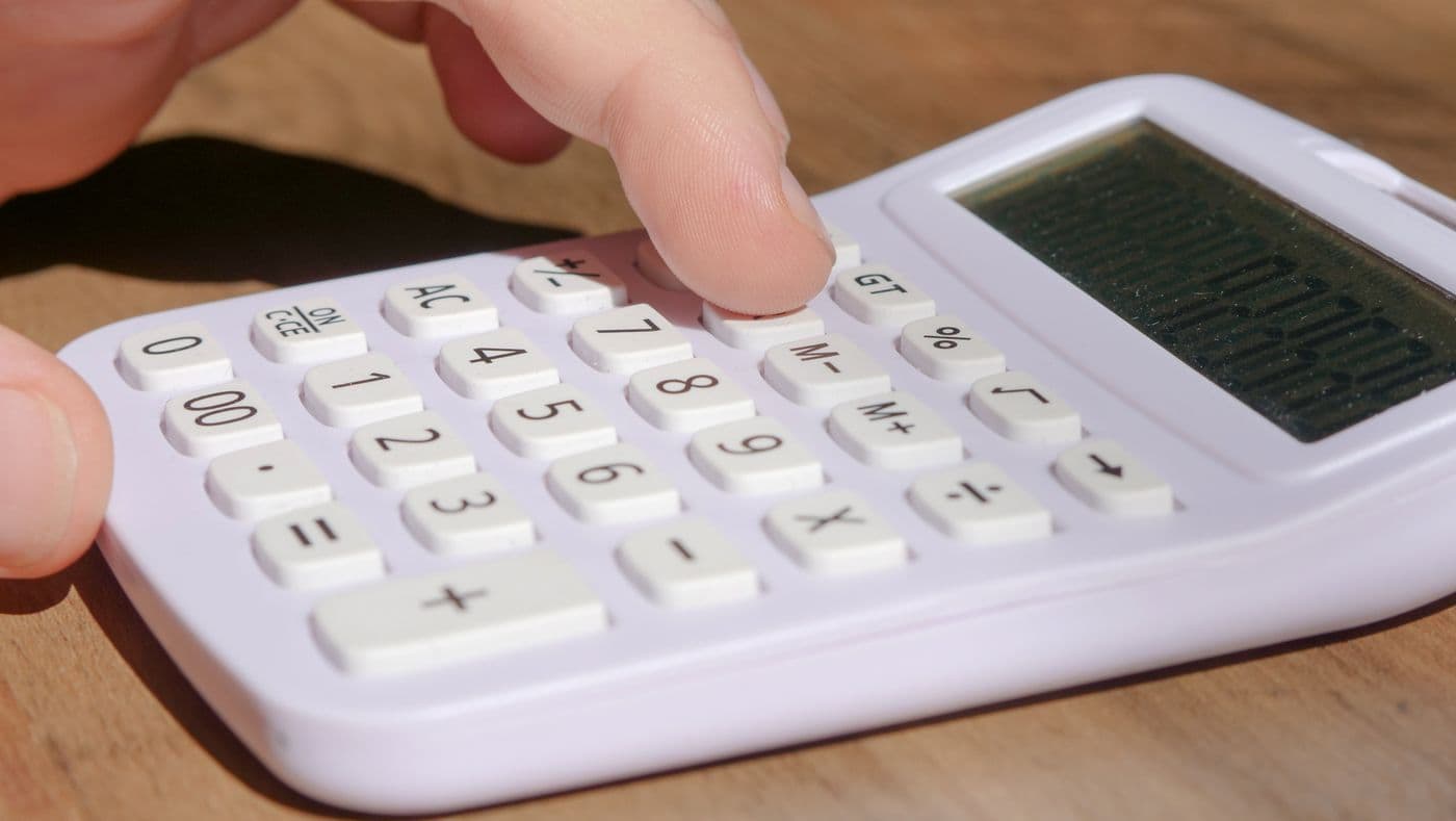 A finger presses the keys of a calculator on a wooden desk.