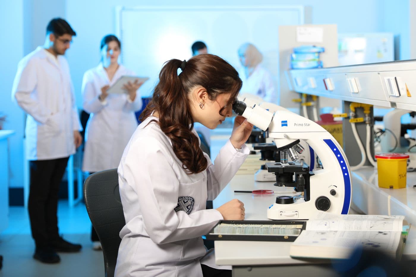 A researcher looks through a microscope in a modern laboratory while colleagues talk behind her.